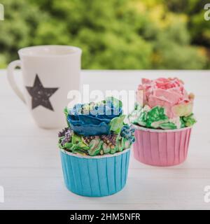 Zwei Muffins mit bunten Butterblumen mit weißer Tasse mit Stern auf weißem Holztisch mit hellgrünem Hintergrund Stockfoto
