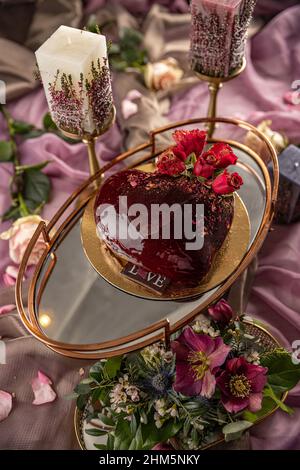 Valentinstag oder Hochzeitstorte Stillleben Konzept. Moderne Mousse Kuchen mit roten Spiegel Glasur in Form eines Herzens mit Rosenkranz verziert Stockfoto