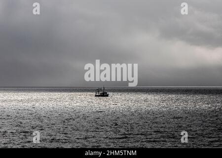 Ein Küstenfischerboot, das an einem bewölkten Tag in der Ardnoquer Bay auf der Kintyre Peninsula, Argyll & Bute, Schottland, in einem Fleck Sonnenlicht gefangen wurde Stockfoto