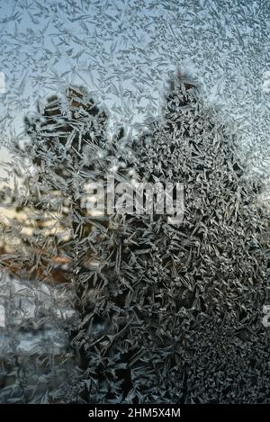 Eiskalte Frost-Kristalle Muster auf Glasfenster des Hauses in der Landschaft, mit Kiefern immergrünen Bäumen draußen während des Sonnenuntergangs, Browntown, Wisconsin, USA. Stockfoto