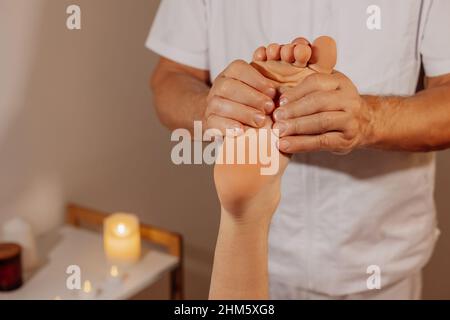 Professionelle Fußmassage im SPA-Salon auf dem Hintergrund von Kerzen. Echter Masseur Therapeut in weißer Uniform macht manuelle Therapie für Mann Bein. Konzept von Wellness, Körper und Gesundheit. Stockfoto