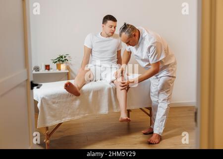 Schöner Masseur Therapeut in weißer Uniform macht manuelle Therapie für junge Sportler Knie. Professionelle Massage-Behandlung und Rehabilitation für Sportler. Konzept von Wellness, Körper und Gesundheit. Stockfoto