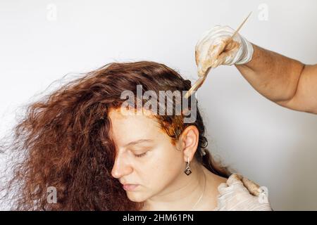 Hände in dünnen Latexhandschuhen, die etwas Färbesubstanz auf Frauenhaare mit weißem Pinsel aus Kunststoff auf weißem Hintergrund auftragen. Haarfärbung zu Hause. Immer Stockfoto