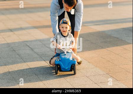 Papa lehrt seinen kleinen Sohn, ein Kinderauto im Park zu fahren, Balance zu halten, Spaß mit der Familie zu haben. Vater und Sohn im Park, der Junge ist drivin Stockfoto