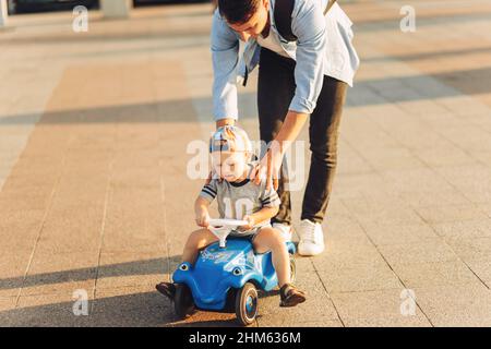 Papa lehrt seinen kleinen Sohn, ein Kinderauto im Park zu fahren, Balance zu halten, Spaß mit der Familie zu haben. Vater und Sohn im Park, der Junge ist drivin Stockfoto