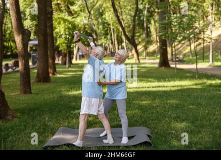 Lächelnde blonde Frau hilft älteren Mann, Sport-Übungen mit Bar im grünen Park zu tun Stockfoto