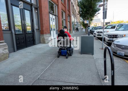 Tacoma, WA USA - ca. August 2021: Mann mittleren Alters in einem motorisierten Rollstuhl, der den Bürgersteig in der Innenstadt entlang geht. Stockfoto