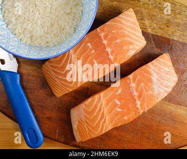 Zwei Stücke frisches Lachsfilet auf einem Schneidebrett mit Messer und ungekochten Reis Stockfoto