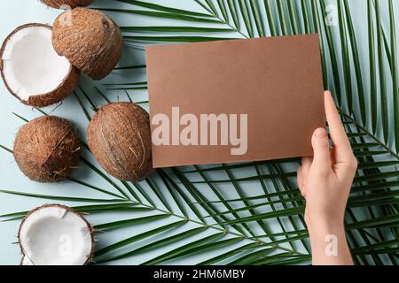 Weibliche Hand mit leerer Karte, Kokosnüssen und Palmenblättern auf farbigem Hintergrund Stockfoto