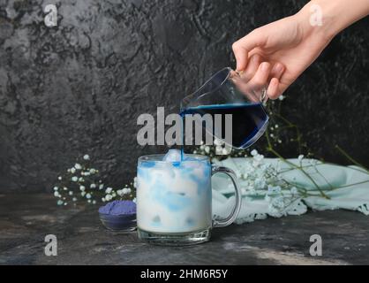 Frau gießt blauen Matcha Latte in die Tasse mit Milch auf dunklem Hintergrund Stockfoto