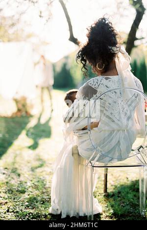 Eine Frau mit einem Baby in den Armen sitzt auf einem Stuhl in einem grünen Garten Stockfoto