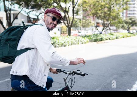 Junger Mann in Sonnenbrille und mit Rucksack auf einem Fahrrad winkend und lächelnd Blick auf die Kamera Stockfoto