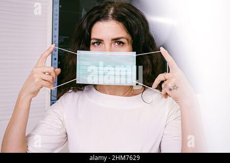 Nahaufnahme einer brünetten Frau in Weiß, die eine medizinische Maske über ihrem Gesicht hält und auf die Kamera blickt. Stockfoto