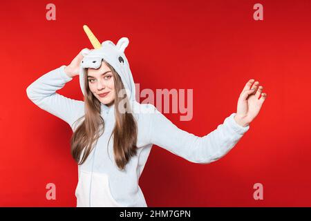 Mädchen im Einhorn-Schlafanzug auf rotem Hintergrund. Übernachtung. Zeit zum Schlafen. Pyjama-Party. Einhorn Kostüm. Stockfoto