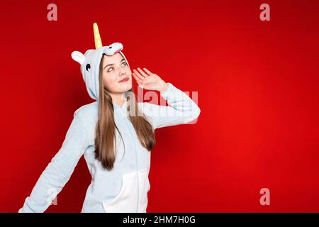 Mädchen im Einhorn-Schlafanzug auf rotem Hintergrund. Übernachtung. Zeit zum Schlafen. Pyjama-Party. Einhorn Kostüm. Herumzutäuschen. Stockfoto