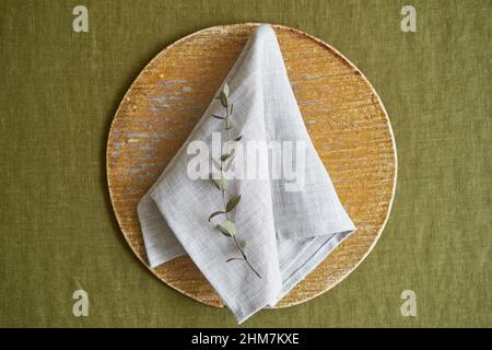 Option für Tabelleneinstellungen. Eine leinengraue Serviette ist ordentlich auf einem braunen Teller gefaltet. Olivfarbener Hintergrund. Stockfoto