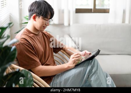 Technologiekonzept der junge Mann, der auf dem Stuhl sitzt und sein iPad hält und etwas darauf notiert. Stockfoto