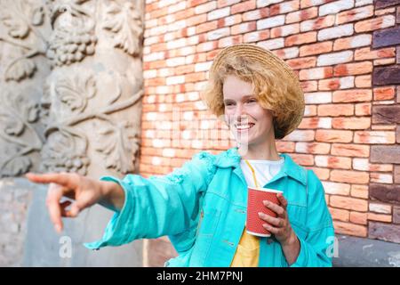 Junge, fröhliche, lockige Rothaarige Frau in blauer Jeansjacke und Strohhut (Reisende), die eine Tasse Kaffee hält und von Sehenswürdigkeiten überrascht ist. Spaß, Sommer, Reisen Stockfoto