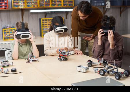 Porträt eines männlichen Lehrers, der einer Gruppe von Kindern hilft, die in der Schule VR-Ausrüstung tragen und Augmented Reality in einem innovativen Roboterkurs einsetzen Stockfoto