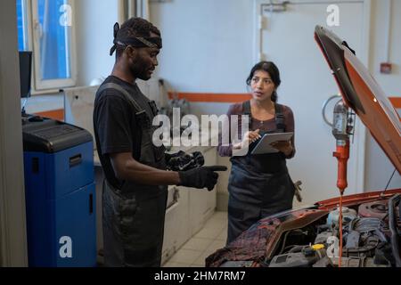 Seitenansicht Porträt von zwei Mechanikern, die das Auto in einem Autogeschäft reparieren, mit Fokus auf afroamerikanischen Arbeitern, die Motor und Kopieraum inspizieren Stockfoto