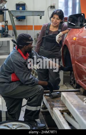 Vertikales Porträt von zwei ethnischen Mechanikern, die das Auto in einer Garage reparieren, und Fokus auf lächelnde junge Frau, die Arbeitskleidung trägt Stockfoto