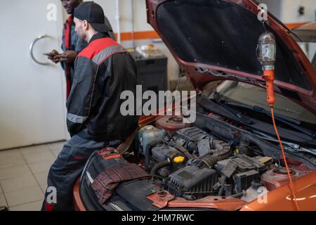 Rückansicht des männlichen Mechanikers, der im Autogeschäft Pause macht, mit offener Motorhaube und Motor im Vordergrund, Kopierraum Stockfoto
