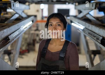Vorderansicht Porträt einer Mechanikerin, die die Kamera anschaut, während sie das Auto auf dem Aufzug in der Autowerkstatt inspiziert, Platz kopieren Stockfoto