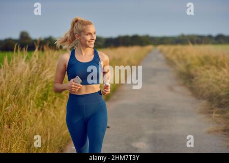 Fit gesunde junge Frau genießen ein Joggen entlang einer Landstraße vorbei an der Kamera mit einem glücklichen Lächeln voller Vitalität in einem aktiven Lifestyle-Konzept Stockfoto