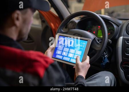 Nahaufnahme des Kfz-Mechanikers mit digitalem Tablet mit Service- und Wartungs-App auf dem Bildschirm, während das Fahrzeug in der Autowerkstatt inspiziert wird Stockfoto