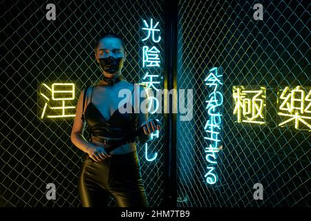 Cyberpunk-Mädchen in Lederkleidung, die mit Fledermaus gegen die Neon-Hieroglyphen steht, was Nirvana und westliches Paradies bedeutet Stockfoto