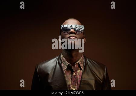 Porträt eines afrikanischen jungen Mannes in stilvoller Lederjacke und Sonnenbrille auf braunem Hintergrund Stockfoto