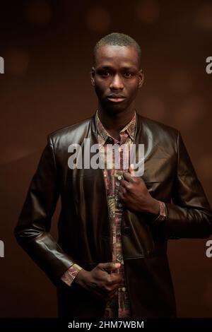 Porträt eines schönen afrikanischen Mannes in Lederjacke, der vor dem braunen Hintergrund auf die Kamera schaut Stockfoto