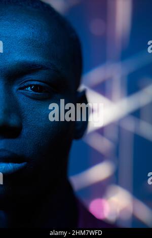 Nahaufnahme eines afrikanischen jungen Mannes, der in einem dunklen Studio mit blauem Licht die Kamera mit unbeschriftem Ausdruck ansieht Stockfoto