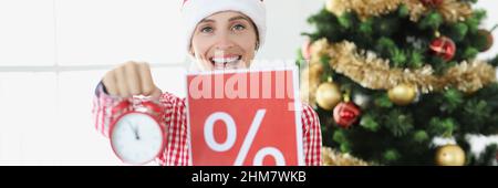 Junge lächelnde Frau im weihnachtsmannhut hält Wecker und Plakat mit Prozent auf dem Hintergrund des Neujahrsbaums Stockfoto