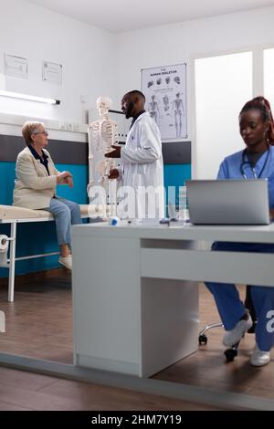 Fachphysiotherapeut Mann zeigt menschlichen Skelettarm erklärt Knochen Krankheit ältere Frau Patientin. Arzt, der während eines Osteoporose-Termins im Krankenhausbüro bei einer medikamentösen Behandlung arbeitet. Stockfoto