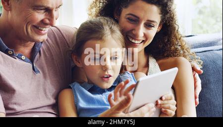 Die Zeit mit der Familie zu einem interaktiven Erlebnis machen. Aufnahme eines niedlichen kleinen Mädchens, das mit ihrer Mutter und ihrem Vater ein digitales Tablet auf dem Sofa zu Hause benutzt. Stockfoto