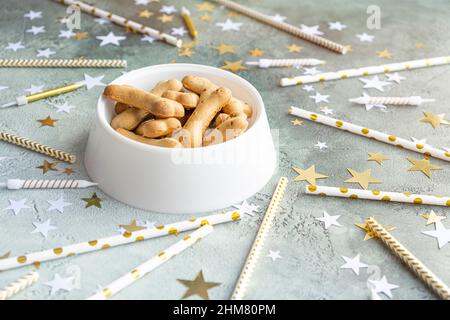 Hundegeburtstag-Leckerbissen in weißer Schale mit Knochengebäck auf festlichem grünen Hintergrund mit goldenen und weißen Sternen, Kerzen und Cocktail-Trinkhalmen. Essen, Genuss Stockfoto