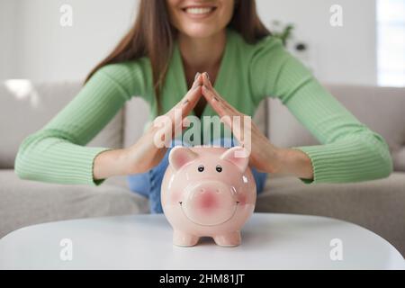 Frau Hände Falten Dach über Sparschwein, was ein Zeichen für die Versicherung ihres Geldes ist. Stockfoto