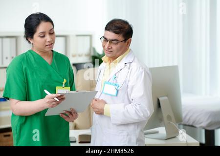 Fokussierter reifer Arzt und Krankenschwester, die auf den Bildschirm des Tablet-Computers schauen und über die Anamnese des Patienten sprechen Stockfoto