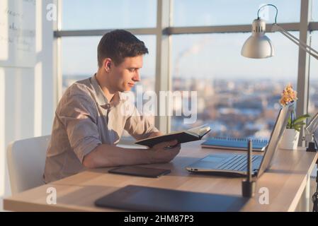 Eine Schriftstellerin, die in einem modernen, hellen Büro arbeitet, neue Ideen in ihr Notebook schreibt, Informationen mithilfe eines tragbaren Computers am Arbeitsplatz sucht Stockfoto