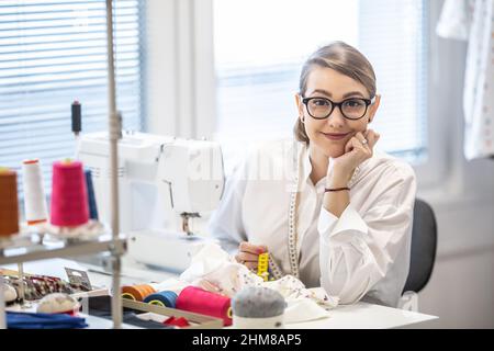 Lächelnder Schneider schaut in die Kamera, die hinter der Nähmaschine sitzt. Stockfoto