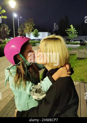 Mutter küsst ihre Tochter in einem rosa Schutzhelm, die gerade gelernt hat, wie sie mit ihren Rollen fährt Stockfoto