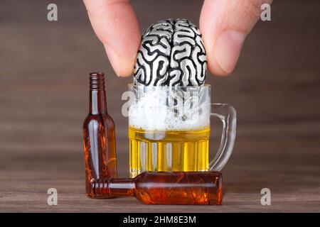Finger ertrinken eine anatomische Miniatur-Kopie des menschlichen Gehirns in einem kleinen Bierkrug vor einem hölzernen Hintergrund. Verlust und Trauer führen zu Substanz Stockfoto