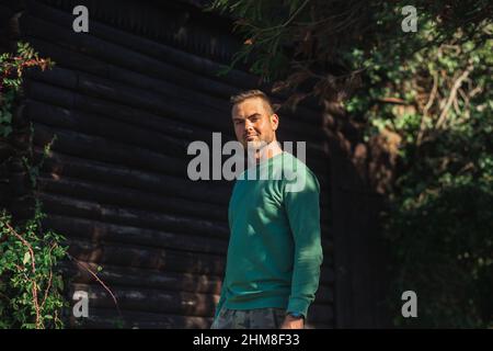 Schöner bärtiger Mann in einem grünen Pullover, der bei Sonnenuntergang an einer Blockwand und grünen Bäumen posiert. Junger umweltfreundlicher Hipster Stockfoto