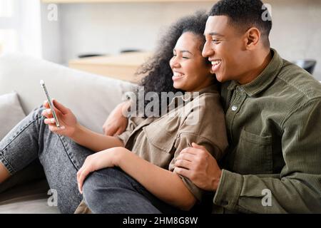 Seitenansicht des verliebten Paares sitzen zu Hause auf der Couch und nutzen Smartphone für lustige Filme. Zeit zu Hause verbringen Konzept Stockfoto