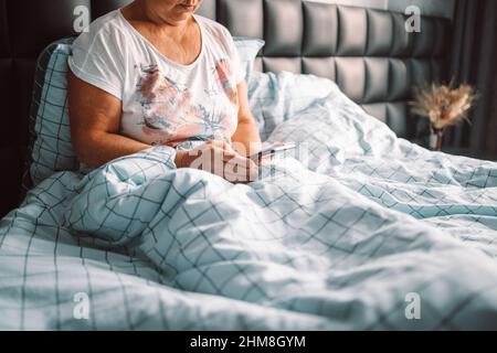 Die blonde Frau mittleren Alters benutzt das Telefon, das im Schlafzimmer auf dem Bett sitzt. Konzept „Guten Morgen, wach auf“ Stockfoto