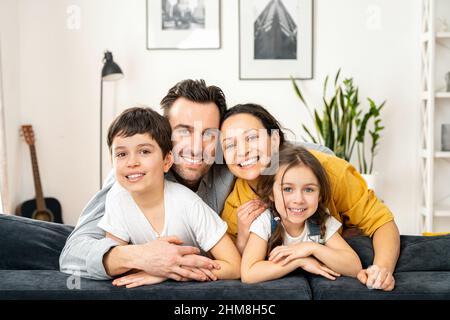 Klassisches Porträt einer glücklichen, lieblichen vierköpfigen Familie, die auf dem Sofa im gemütlichen Wohnzimmer sitzt. Schöne Mutter, hübscher Vater, lustige Kleinkind-Tochter und Sohn im Schulalter, der die Kamera anschaut und fröhlich lächelt Stockfoto