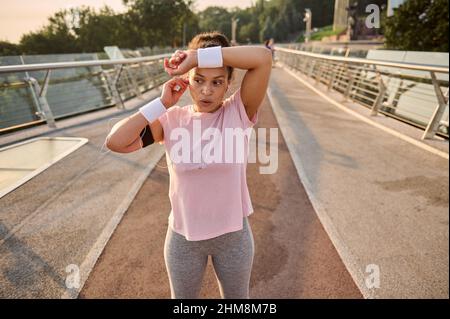 Erschöpfte sportliche Läuferin, Sportlerin, die auf einem Laufband steht und nach einem morgendlichen Joggen in der Stadt mit einem weißen Frottee-Armband schwitzt Stockfoto