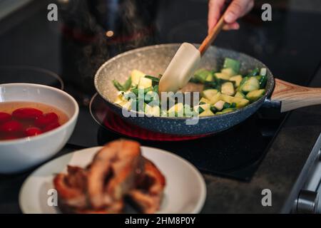 Nahaufnahme einer unkenntlichen Frau, die das Mittagessen in der Küche zubereitet. Gegrilltes Forellensteak. Braten unter Rühren. Die Zucchini und die grünen Bohnen werden in der Pfanne gebraten. Strom Stockfoto