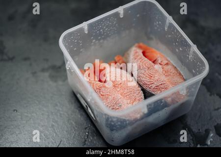 Blick von oben auf frisch gefrorenen rohen roten Lachsfisch. Gefrorene Lachssteaks in einer Plastikbox oder einem Behälter auf dunkelgrauem Hintergrund. Stockfoto